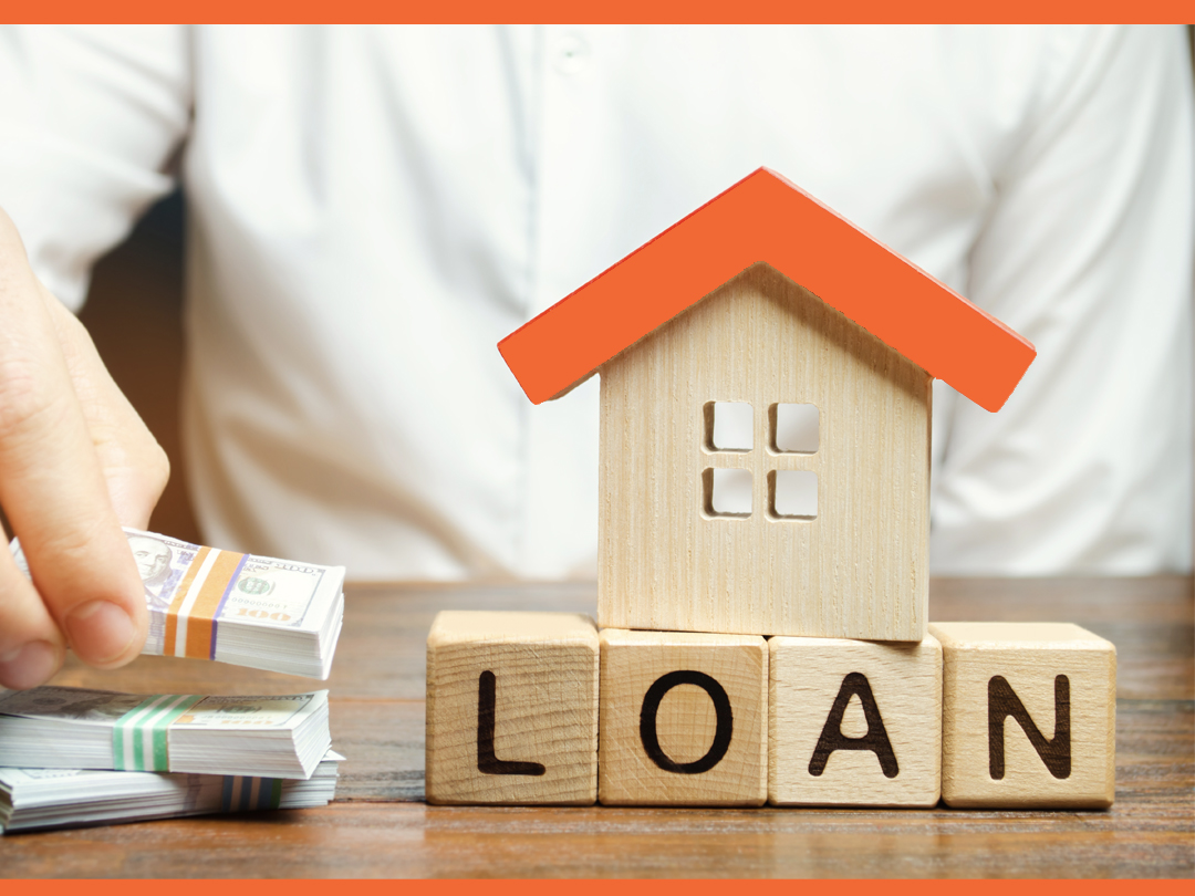 Wooden house sitting on wooden blocks spelling "LOAN" next to a man stacking mini paper money.