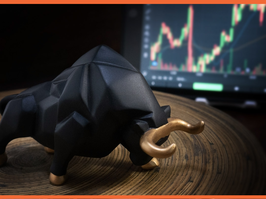 Black metal bull with gold horns sitting in front of a computer screen with economic graph.