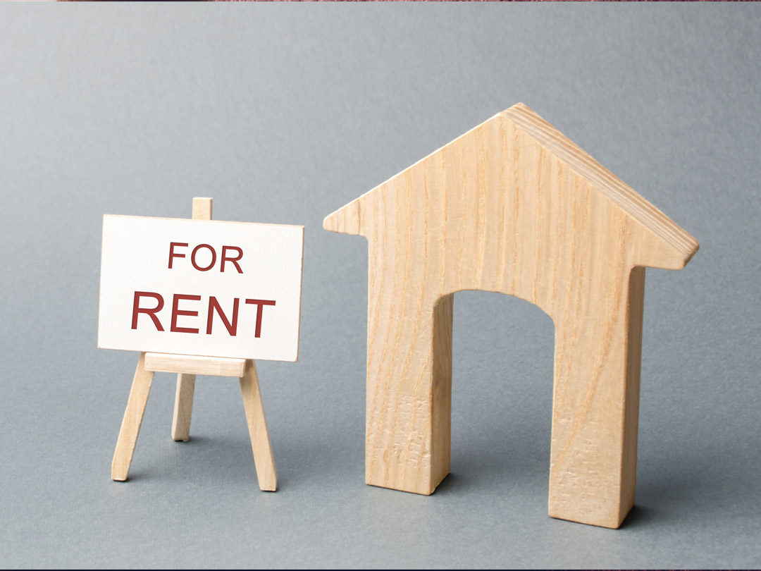 Wooden outline of a house and a sign saying "FOR RENT" laying on a wooden easel on a gray background.