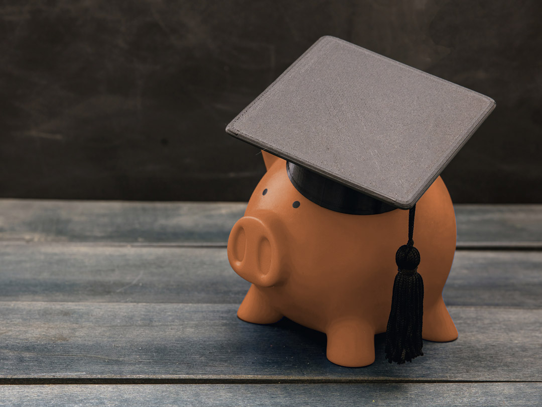 Orange piggy bank wearing a student's graduation hat while resting on a faded gray wooden table.